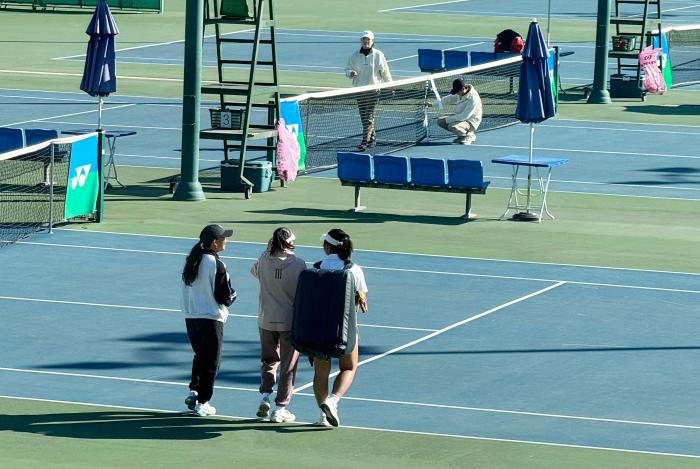 매치 준비를 하고 있는 심판들과 커다란 가방을 메고 코트로 들어서고 있는 주니어 선수들